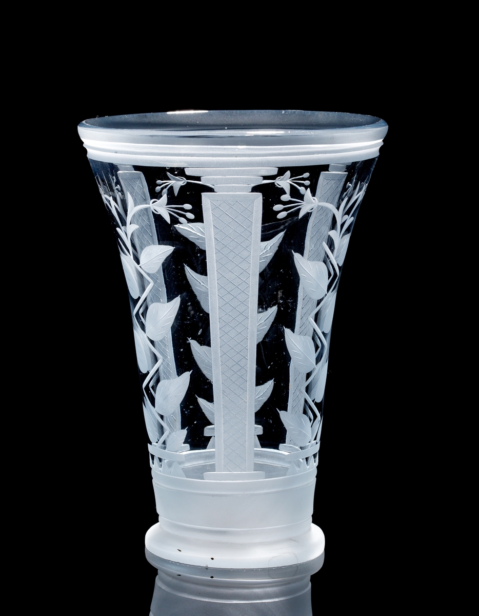 Formgiven av Edward Hald. Ljusblå trumpetformad vas med rund klack. Slipad dekor i form av nätmönstrade pelare och
stiliserade blomrankor.