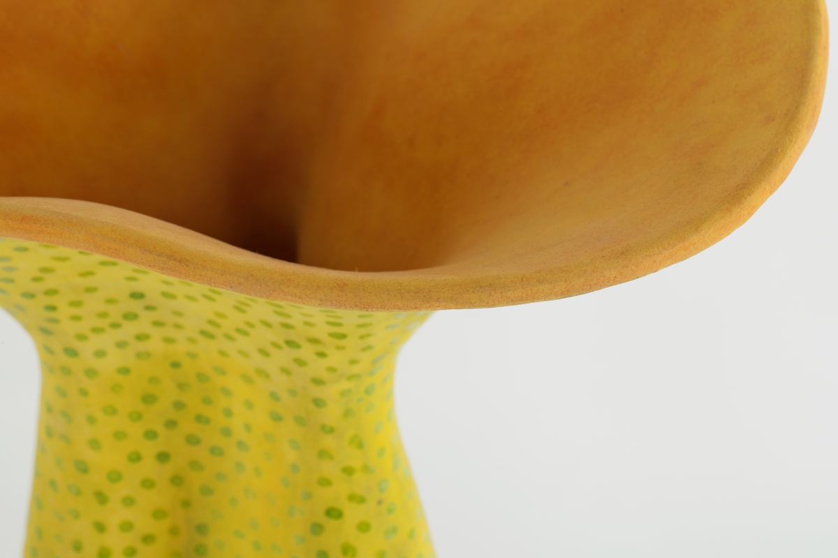 Keramikkvase i begerform som vertikalt svinger ut og inn i buklede, regelmessige kuleformer som bunner i fem dråpeformede "føtter". Munningen åpner seg buktede som en blomsterklokke. Yttersiden er matt gul og innsiden er matt rød-oransje. Bunnens innside er blank svart.