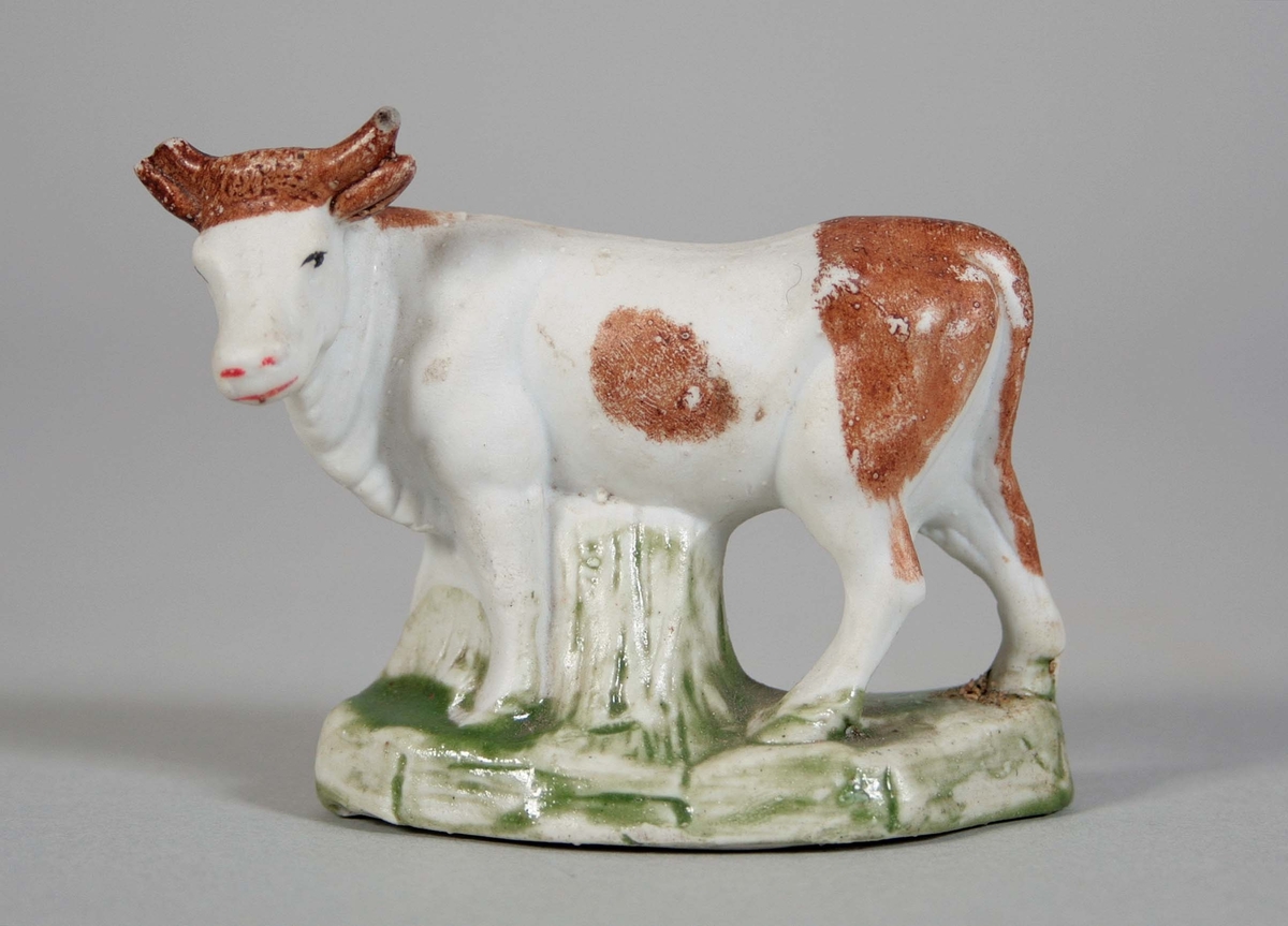 Stående ko, med vänstervridet huvud på oval sockel i oglaserat porslin. Vit med partier i rödbrun färg. Sockeln med fläckar i grönt. Höger horn med nagg.