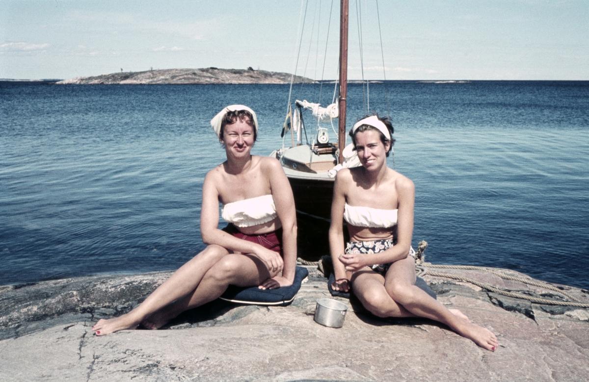 Dagmar Dahl (t v) och Aina Palmér sittande på en klipphäll framför folkbåten F-S133 KARP.