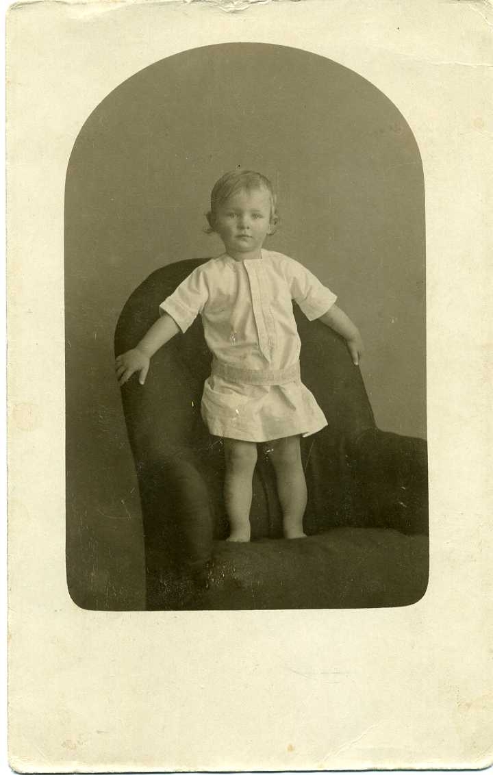 Porträtt av en liten pojke i ljus särk som står i en fåtölj. Vykort.