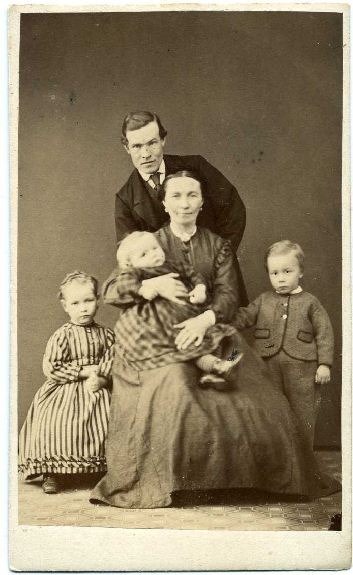 En familjegrupp. En kvinna sitter med ett litet barn i sin famn. En flicka står vill vänster om henne och en pojke till höger. Bakom dem lutar sig en man lätt över dem.