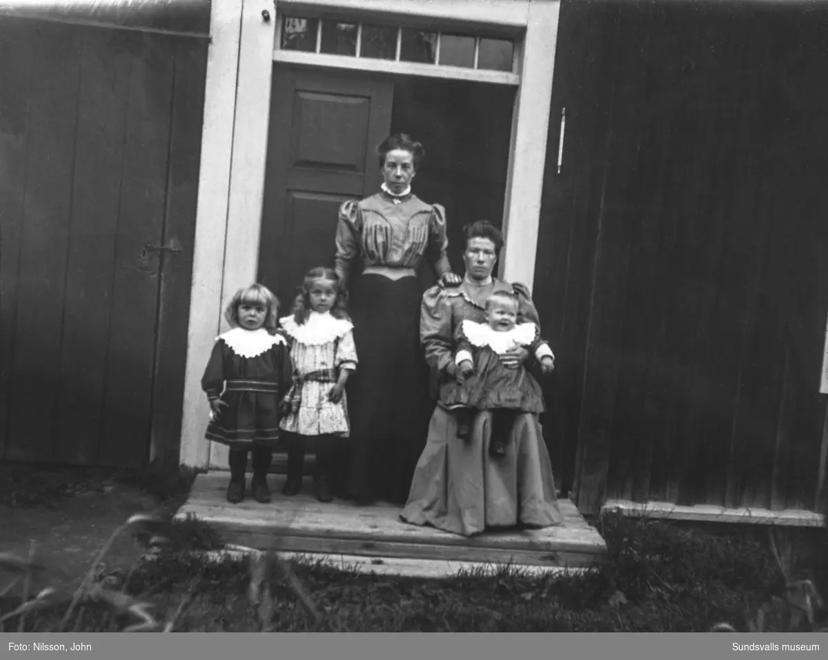 Familjeporträtt med två kvinnor och tre små barn framför en dubbeldörr med överljus. På bild nummer två från samma tillfälle finns även en äldre man med.