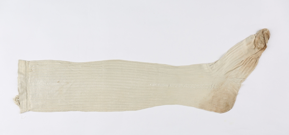 Hvit silketrikot, stripet (finere og grovere masker) med glatt tå og glatt kant oppe. De har søm bak, under foten og tversover ved tåen. Brodert pil.