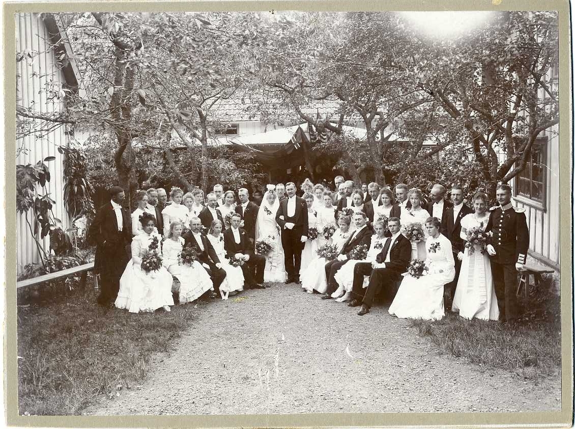 Ett brudpar med gäster står i en trädgård. På farstun bakom dem hänger bland annat en unionsflagga och stjärnbaneret.