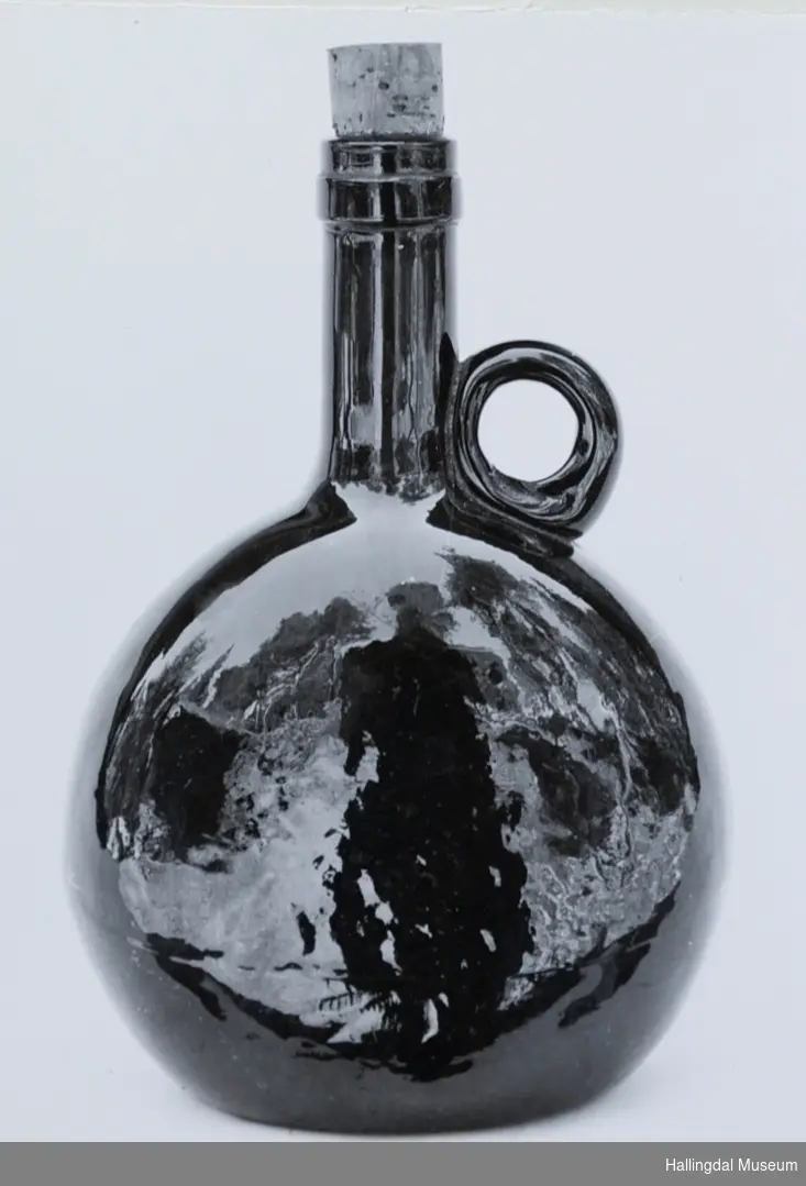 Selve flaske er tilnærmet sirkelrund fra siden, med ovalt tverrsnitt. Tuten sylindrisk. I overgangen til tuten er en rund kork.
6/6-68. Arne L. Christensen.
