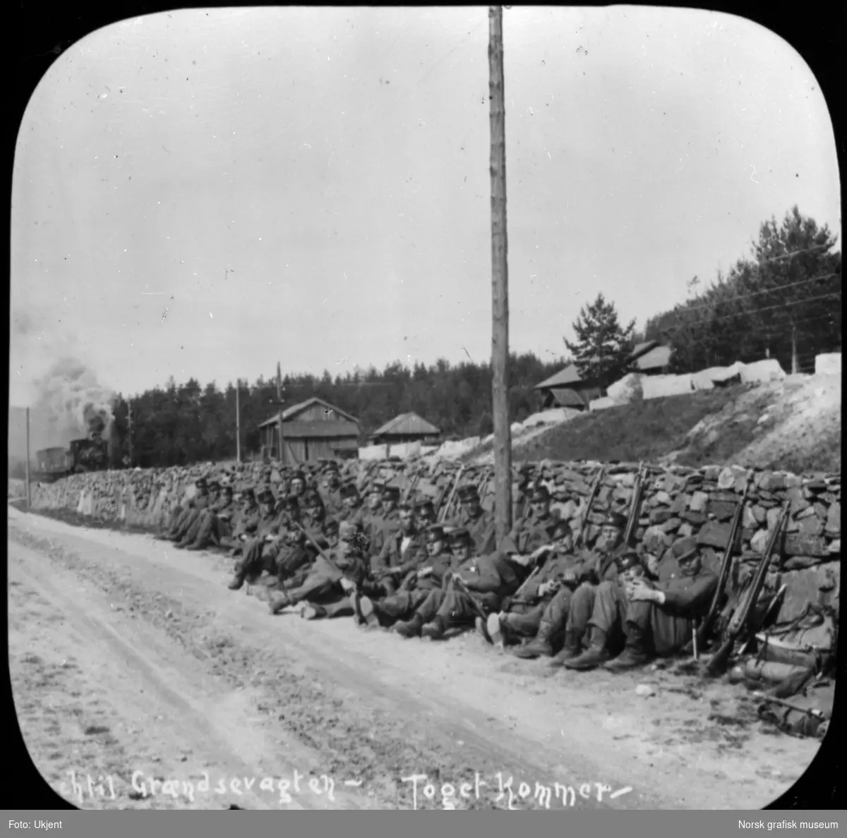 Soldater fra Norske jegerkorps eller grensevakten sitter i en veikant og venter på toget . Et damplokomotiv kommer i bakgrunnen.