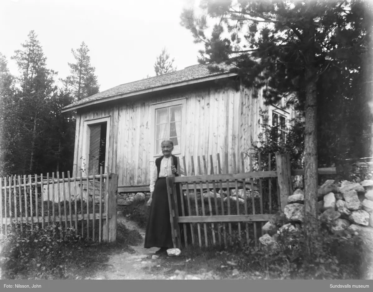 En äldre kvinna står vid staketet framför en liten stuga med omålad träpanel.