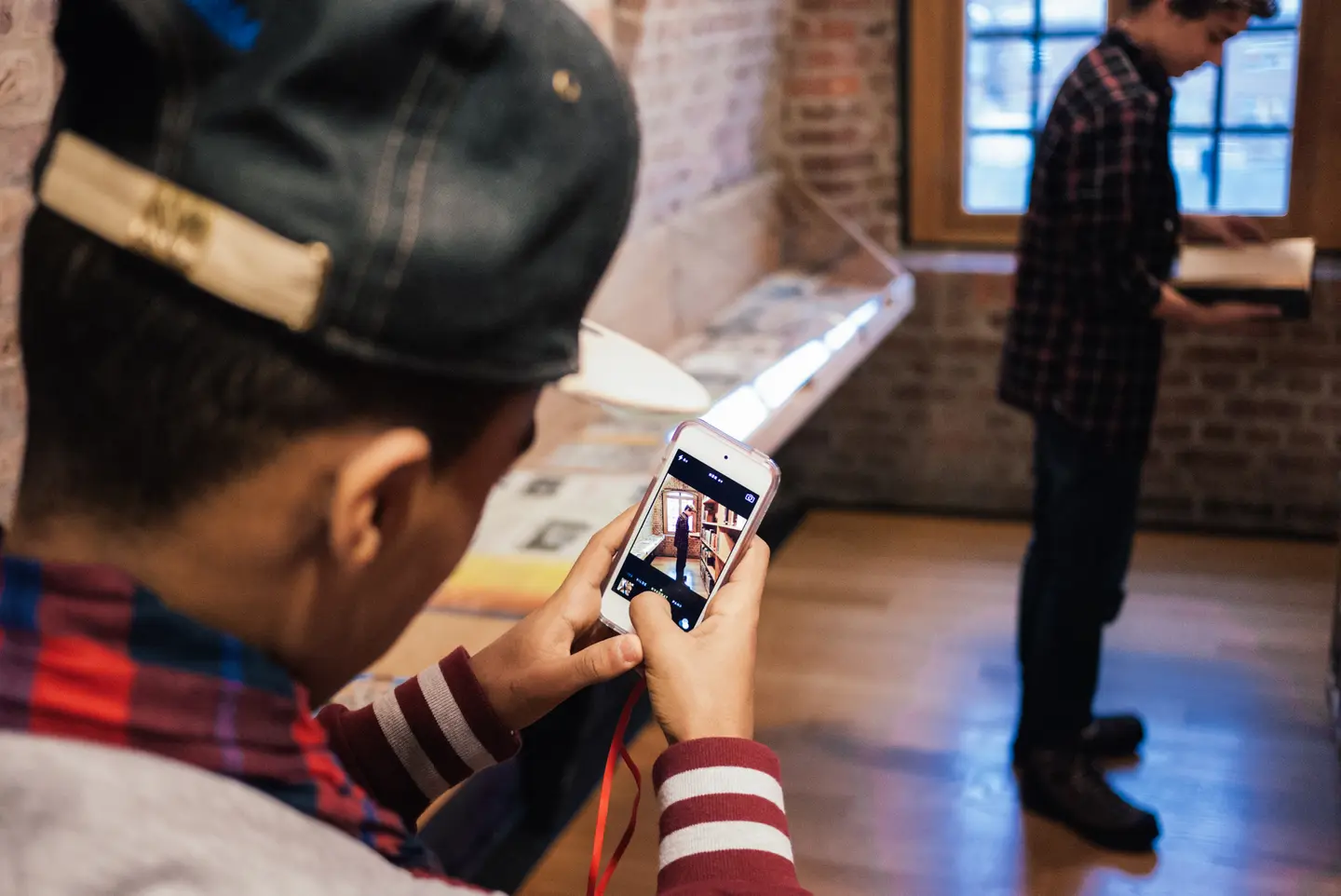 En gutt med caps fotograferer en annen gutt som står og leser i en bok. Gutten fotograferer med mobil, og vi ser bildet på skjermen. Fotografi fra Preus museums bibliotek.