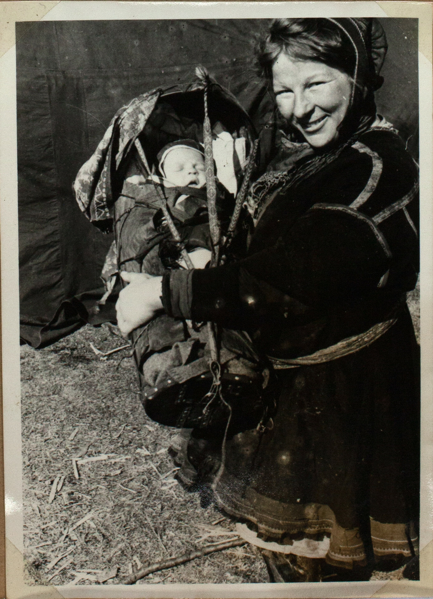 Bildet viser en samisk kvinne som kneler og holder gutten sin i en komse i sin fang. Barnet sover mens hans mor ser inn i kameraet med en stolt smil. Bak dem ser man et telt. 