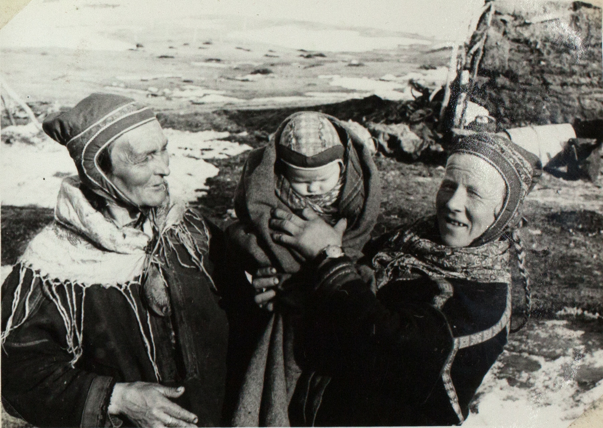 Bildet viser to eldre, samiske kvinner i tradisjonelle antrekk. De holder deres barnebarn opp. En av dem kikker i kameraet, mens den andre ser på gutten. I bakgrunnen ser man en gamme og et delvis snødekket landskap.