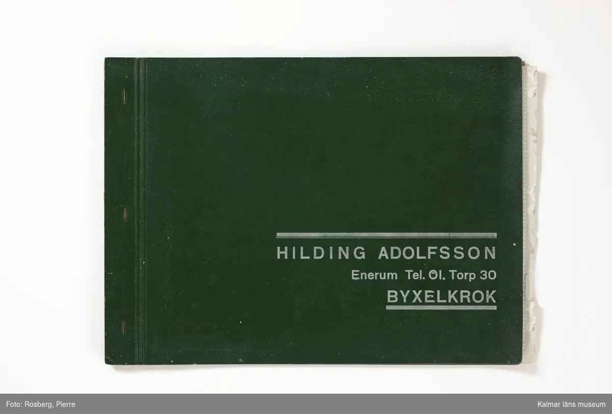 KLM 46599:4. Tapetkatalog. Katalog med pärm av grön papp eller kartong. Bunden med hjälp av tre häftklamrar. Text i silver på pärmens framsida: HILDING ADOLFSSON Enerum Tel. 01.Torp 30 BYXELKROK. Katalogen innehåller plastförstärkta tapetprover tillverkade av papper, med olika strukturer, färger och mönster, moderna under 1950 och 1960-talen. Större delen av tapetproverna saknas och endast fyra återstår, varav två av dessa ligger lösa. Inuti katalogen ett löst blad med prisuppgifter från Emmaboda Tapetfabrik AB, tryckt 1962.