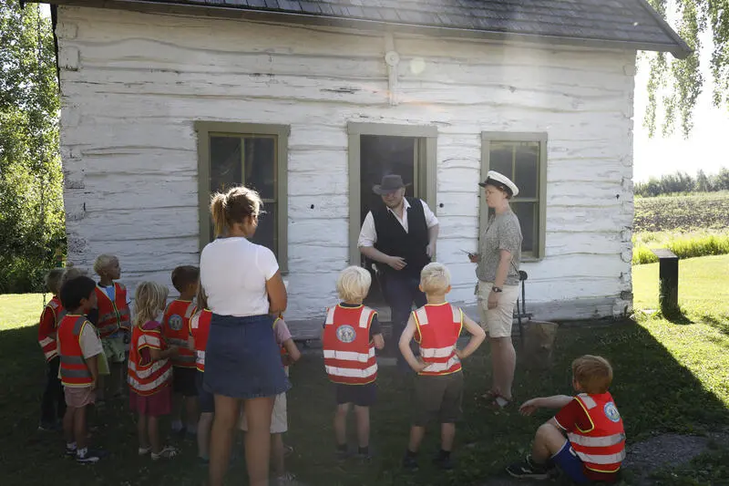 Små barnehagebarn med oransje vester står sammen med en voksen utenfor skolestua på Norsk utvandrermuseum og får høre om historien av en mann i historiske klær.