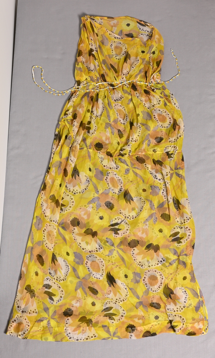 Blommig klänning av sidenchiffong, tryckt mönster i gulgrönt, brunlila, svart och brunt. Rak modell som lämnar vänster axel bar, hög midja. "Skärp" av resårband med fastsydda glaspärlor i ljusblått, ljusgrönt och gulbrunt (BM54231).