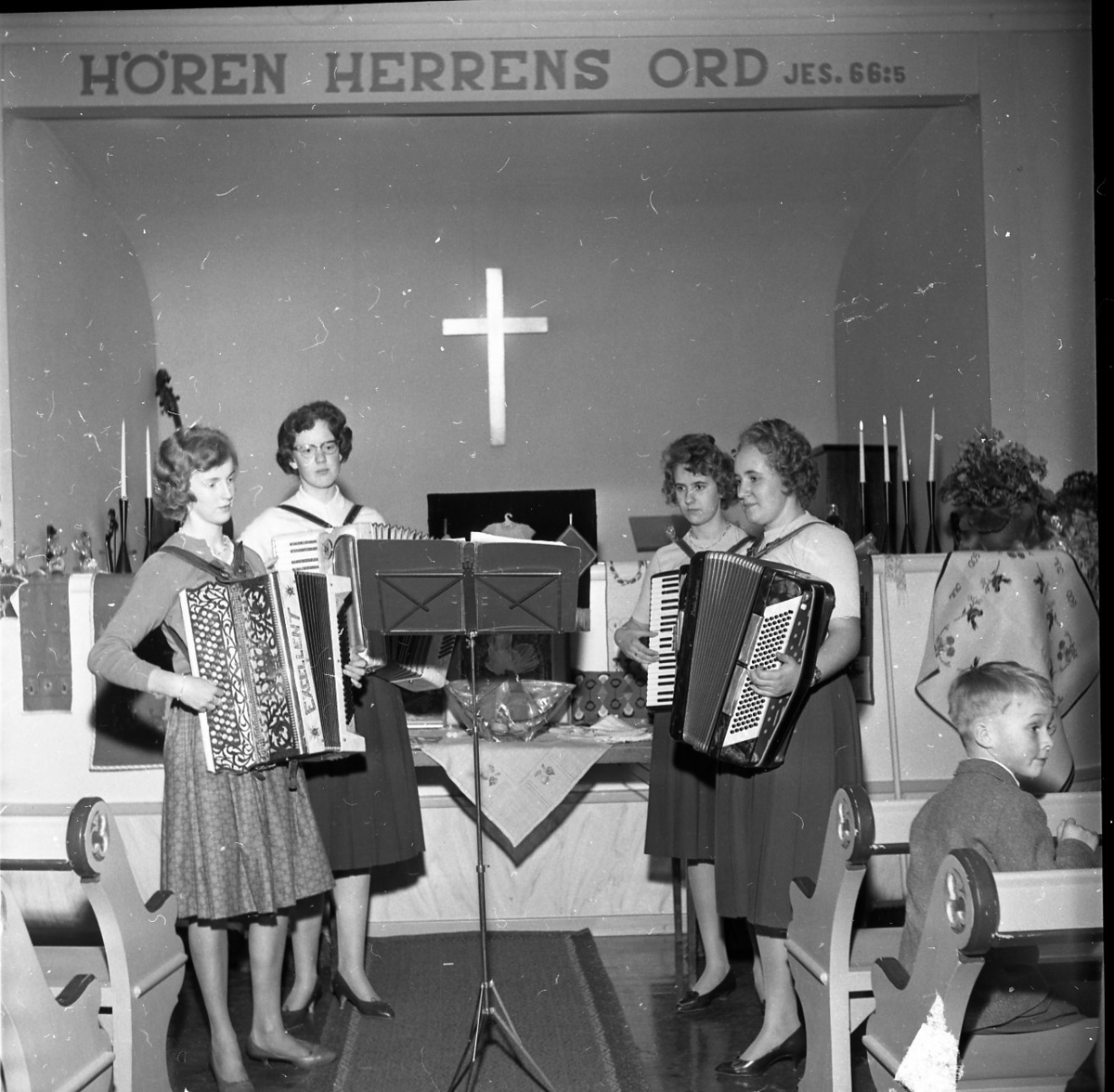 Fyra kvinnor står och spelar dragspel framför ett altare. Bakom dem hänger ett kors. På väggen bakom dem står "HÖREN HERRENS ORD Jes: 66.5". Interiörbild från Norra missionshuset i Skärstad.