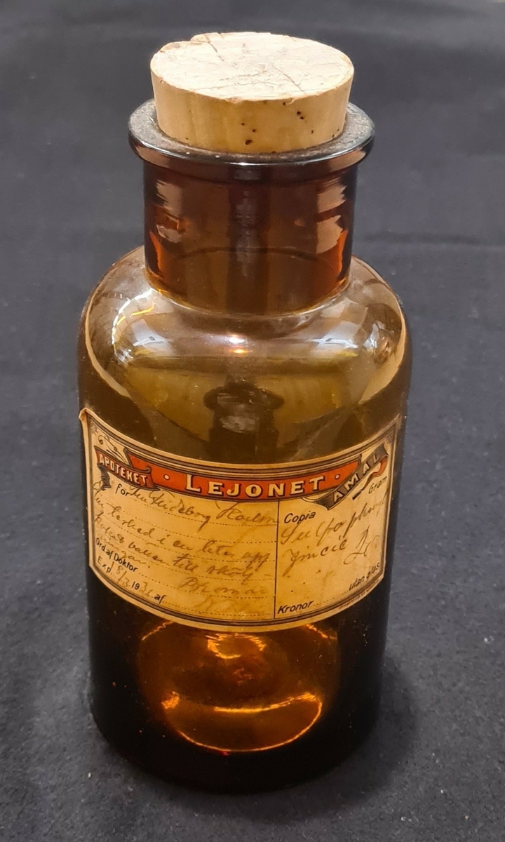 Glasflaska i brunt glas med kork. Vit etikett med handskriven text. Flaskan kommer från Apotek Lejonet i Åmål.