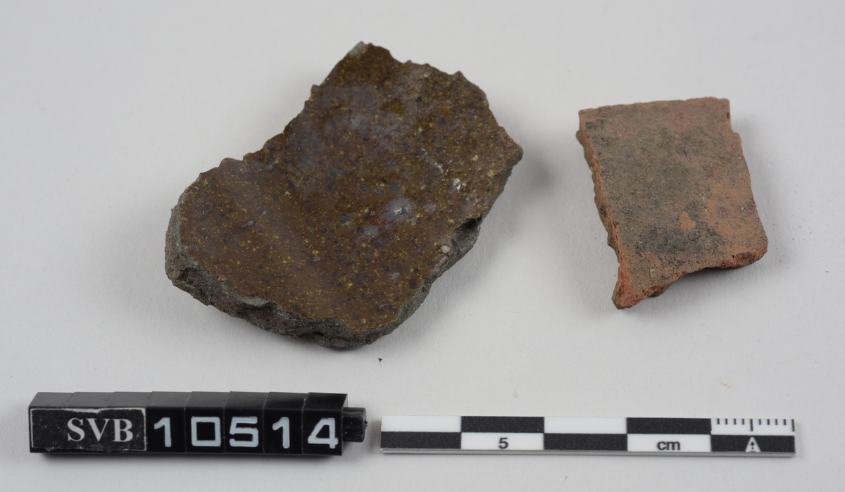 To fragmenter av keramikk. Det største fragmentet er laget av grå leire og har brun glasuer på begge sider. Det andre er laget av rød leire og har brun glasur på innsiden.
