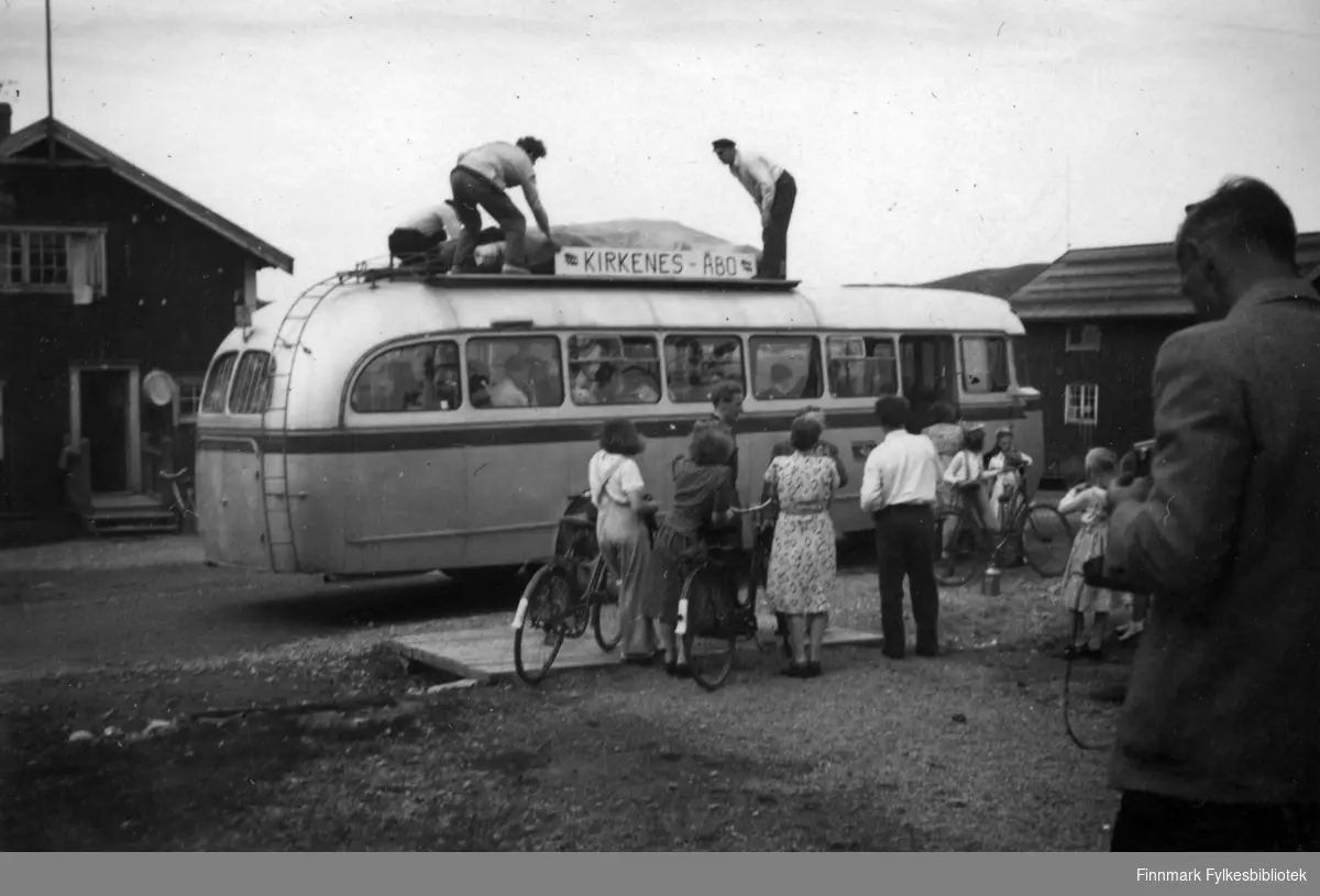 Avreise til AUF-stevnet i Åbo i Finland, 1949. På taket på bussen er det et skilt hvor det står 'Kirkenes - Åbo'. Bildet er tatt i EgneHjemsveien i Bjørnevatn. Flere personer står utenfor bussen, flere med sykler. Noen menn er på taket til bussen