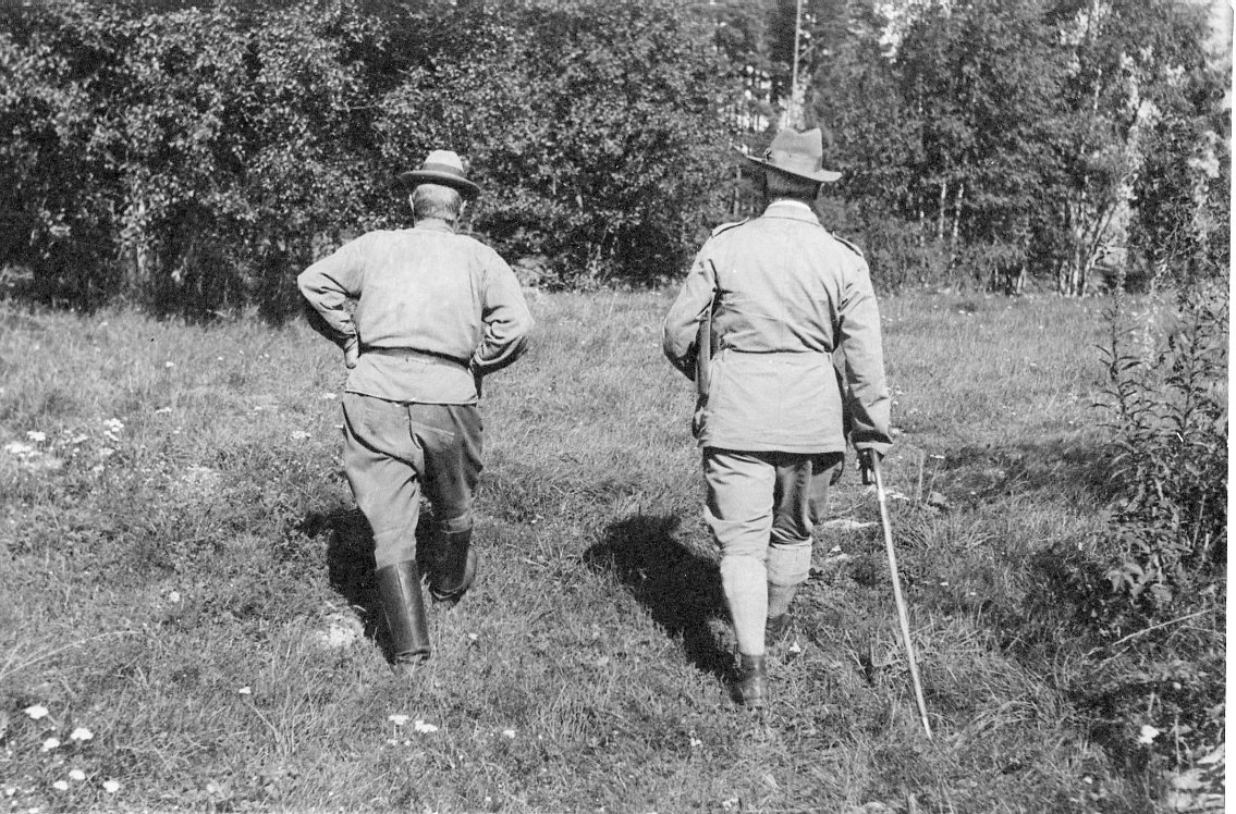 Två män i fältmässig klädsel promenerar på en äng mot ett skogsparti, den ena med en käpp. De är fotograferade bakifrån.