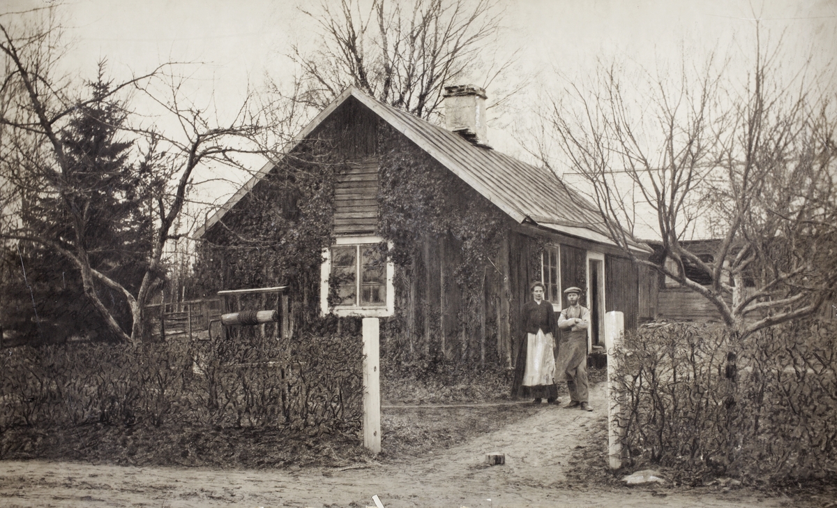 En man och en kvinna står utanför sitt lilla bostadshus. I bakgrunden skymtar en ladugård/lada.
