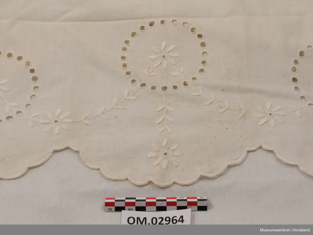 Hvit pyntehåndduk med initialene LM brodert nederst. Øverst er tre ringer formet av sydde hull og brodert blomsterguirlander.