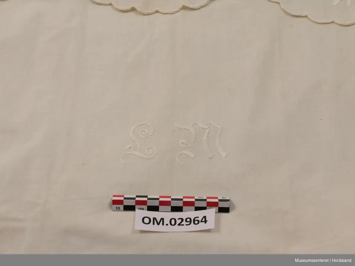 Hvit pyntehåndduk med initialene LM brodert nederst. Øverst er tre ringer formet av sydde hull og brodert blomsterguirlander.