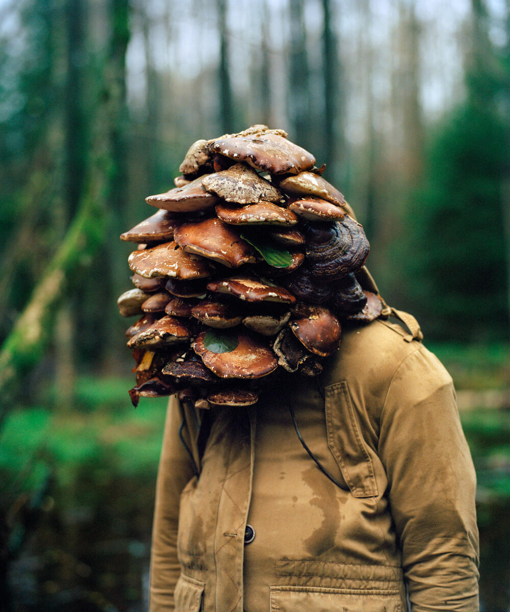 En kvinne står i en skog, hodet hennes er dekket av kjuker, eller sopp som vokser på stammen på trær. Hun har en beige jakke på, og ansiktet er helt skjult av soppen. Fotografi.