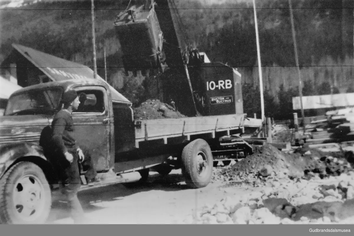 Ei gravemaskin lastar masse frå tomta til Bismo Samvirkelag på ein lastebil. Sevald Forberg, Skogli (f. 1925) er sjåfør