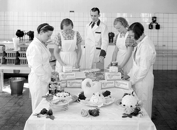 Arbetsklädda kvinnor och man vid bord med glassprodukter.
Fotografens ant: Karlstadsortens - Mejeriförening.