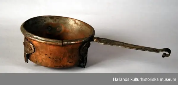 Kastrull av koppar, med skaft och rester av tre avbrutna ben av järn. Rundad botten, nästan cylindriska sidor med rundad mynningskant. Handtaget är platt och avslutat i en krok.