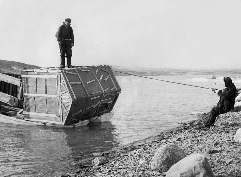 Bildet viser en hytte som ligger på en flåte laget av to båter. Flåten taues til land av en mann.