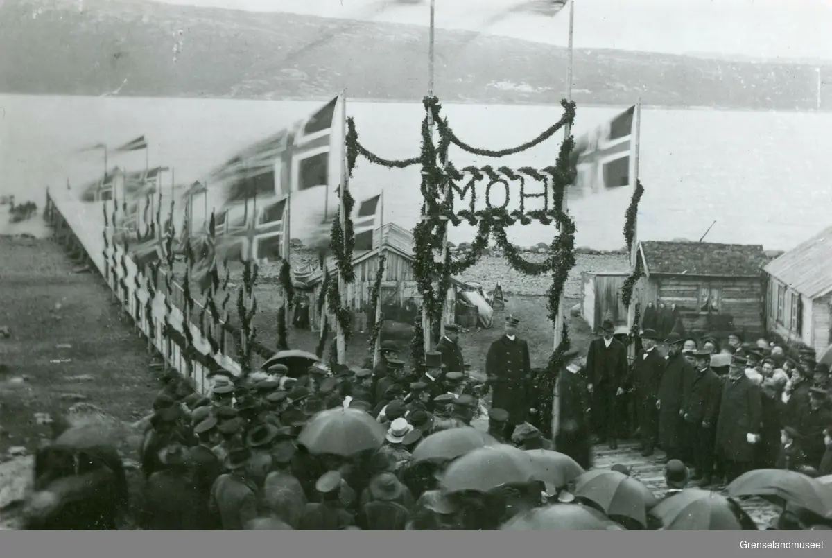 Kongebesøk i 1907. Æresporten. Kong Haakon VII og dronning Mauds besøk i Kirkenes 27.07.1907. Vi ser kaia pyntet med flagg og befolkningen er festkledt i anledningen.