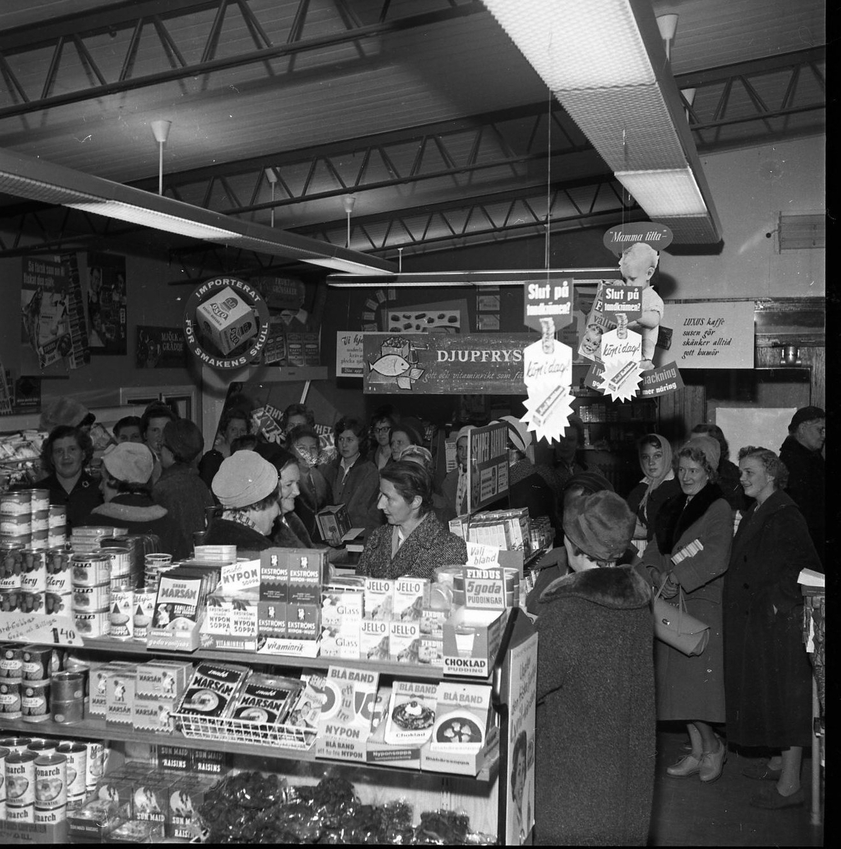Kvinnor står packade i en mataffär. Från taket hänger reklam för mat och tandkräm. På hyllorna står matvaror med varumärken som Ekströms, Blå band och Jell-o.