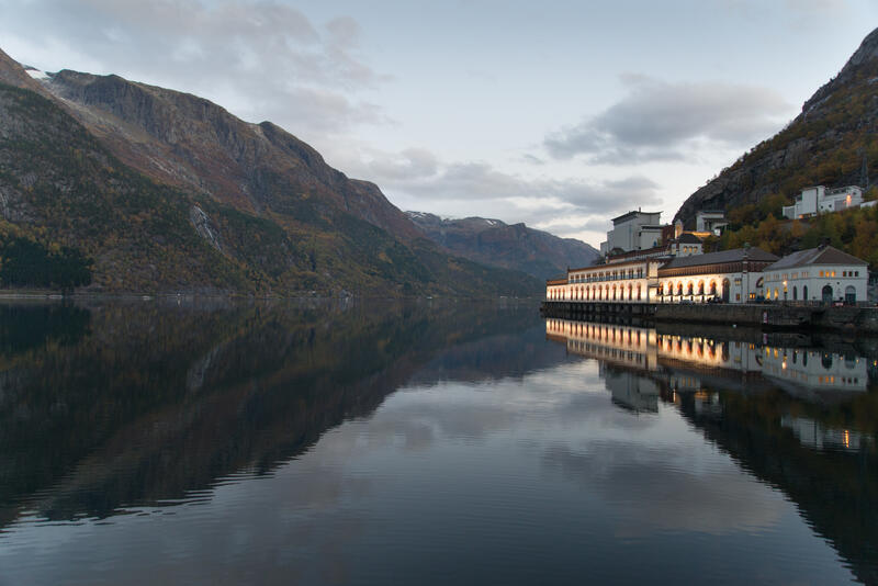 Ein ser fjord, fjell og krafthistorie i vakker harmoni. Kraftstajonen speiler seg i fjorden. Foto.