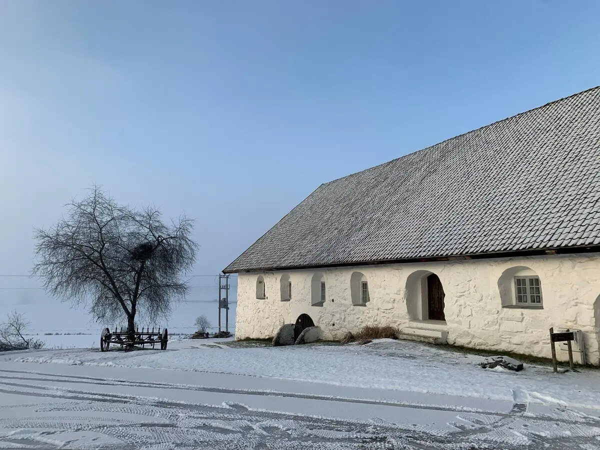 Bildet viser et hvitt steinfjøs med blå himmel. Det er hvit snø foran fjøset med tydelige hjulspor etter biler.