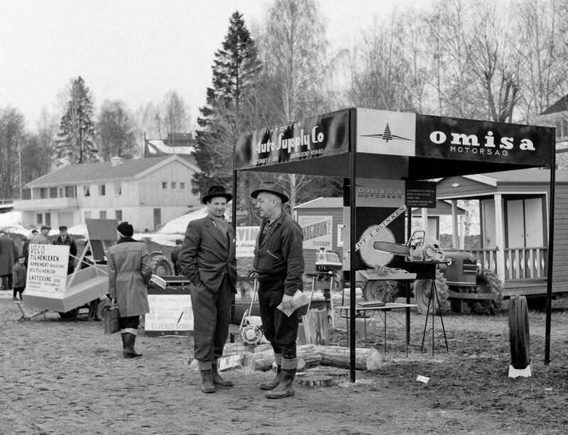 To menn står foran en salgsbod hvor det er utstilt motorsag. Bildet er i svarthvit og er tatt på 60 tallet
