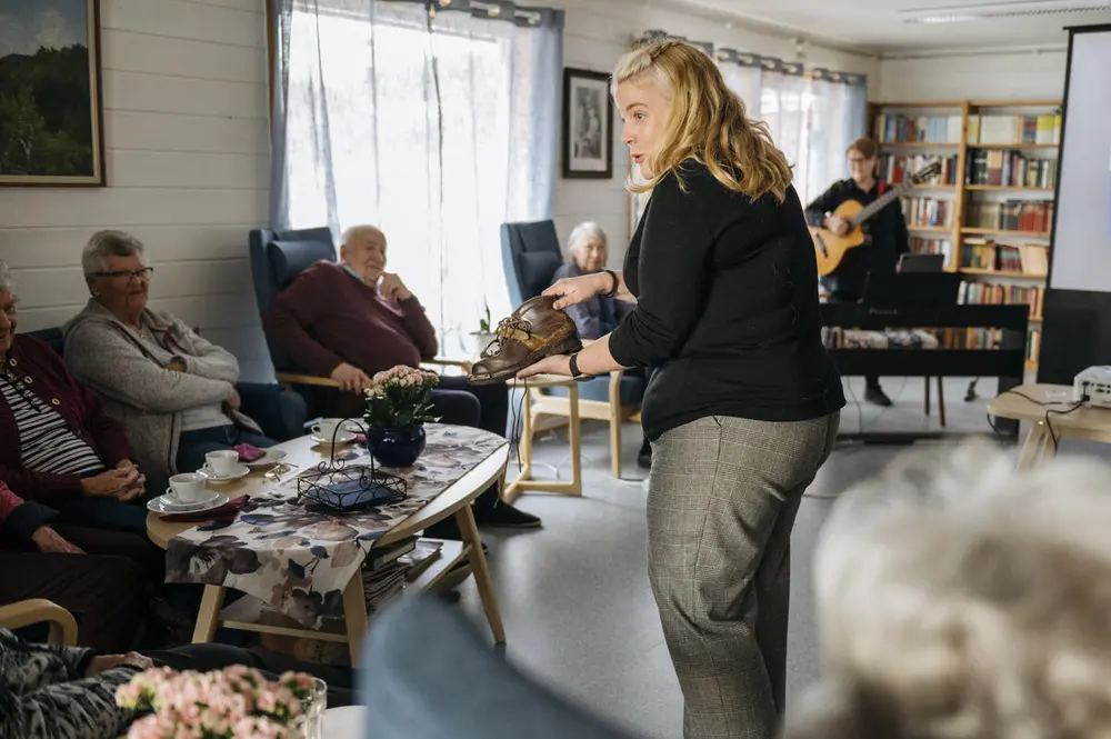 En dame forteller foran flere lyttere. Lytterne er eldre og sitter rundt og hører på. I bakgrunn skimtes en person med gitar