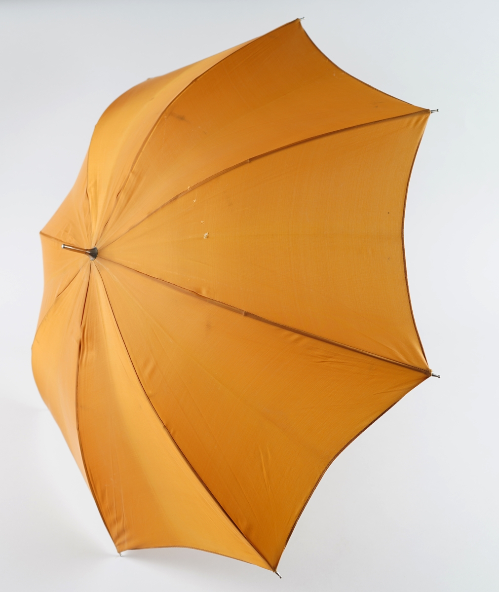 Brun paraply med handtak i svart (syntetisk?) skinn. Trykk-knapp med logo danna av to paraply-symbol. Manuell mekanisme til å slå opp paraplyen.