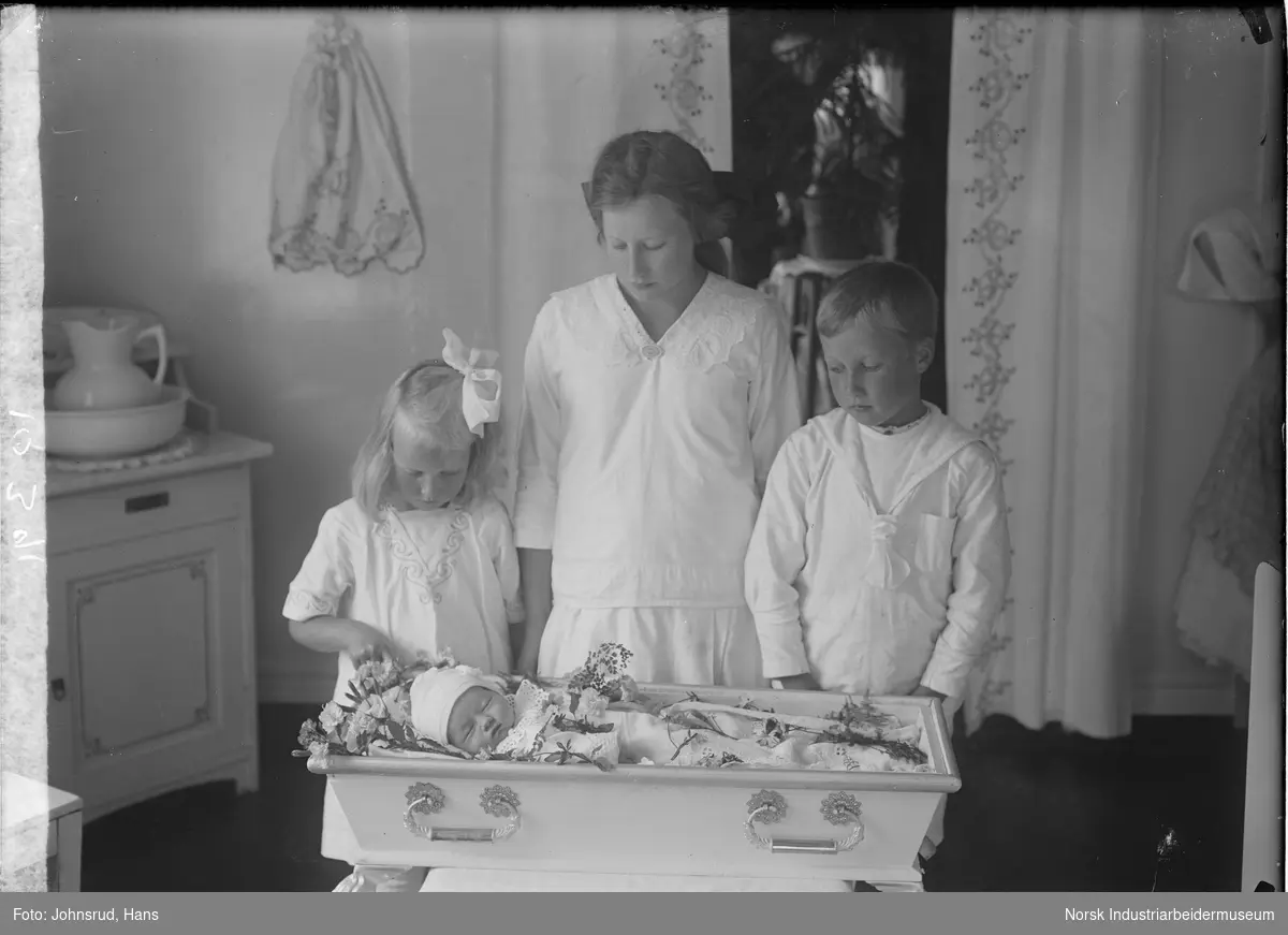 Port mortem fotografi. To jenter og en gutt stående ved åpen barnekiste. Spedbarnet i kisten har blomster liggende rundt seg.