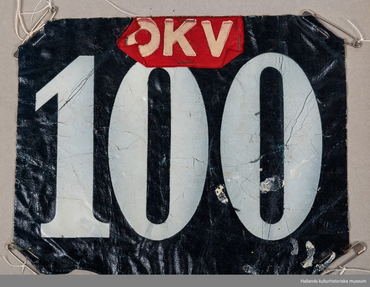 Nummerlappar av galonväv, svart botten med vita siffror: 100. Påsatt bit av rött sidenband med bokstäverna "OKV". Säkerhetsnålar i hörnen för att fästa på kläderna.