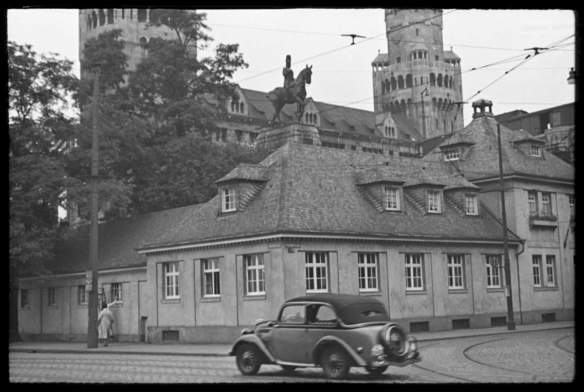 Ved Hohenzollernbrücke i Köln, på nord-vestsiden like ved Köln-domen. Gate med trikkespor, delvis skjult bak bygninger er bruas tårn og portal brua. Rytterstatuen framstiller Fredrik Vilhelm 3., som var konge av Preussen fra 1797 til 1840. Bilen er en Ford Eifel 1937–1938.
