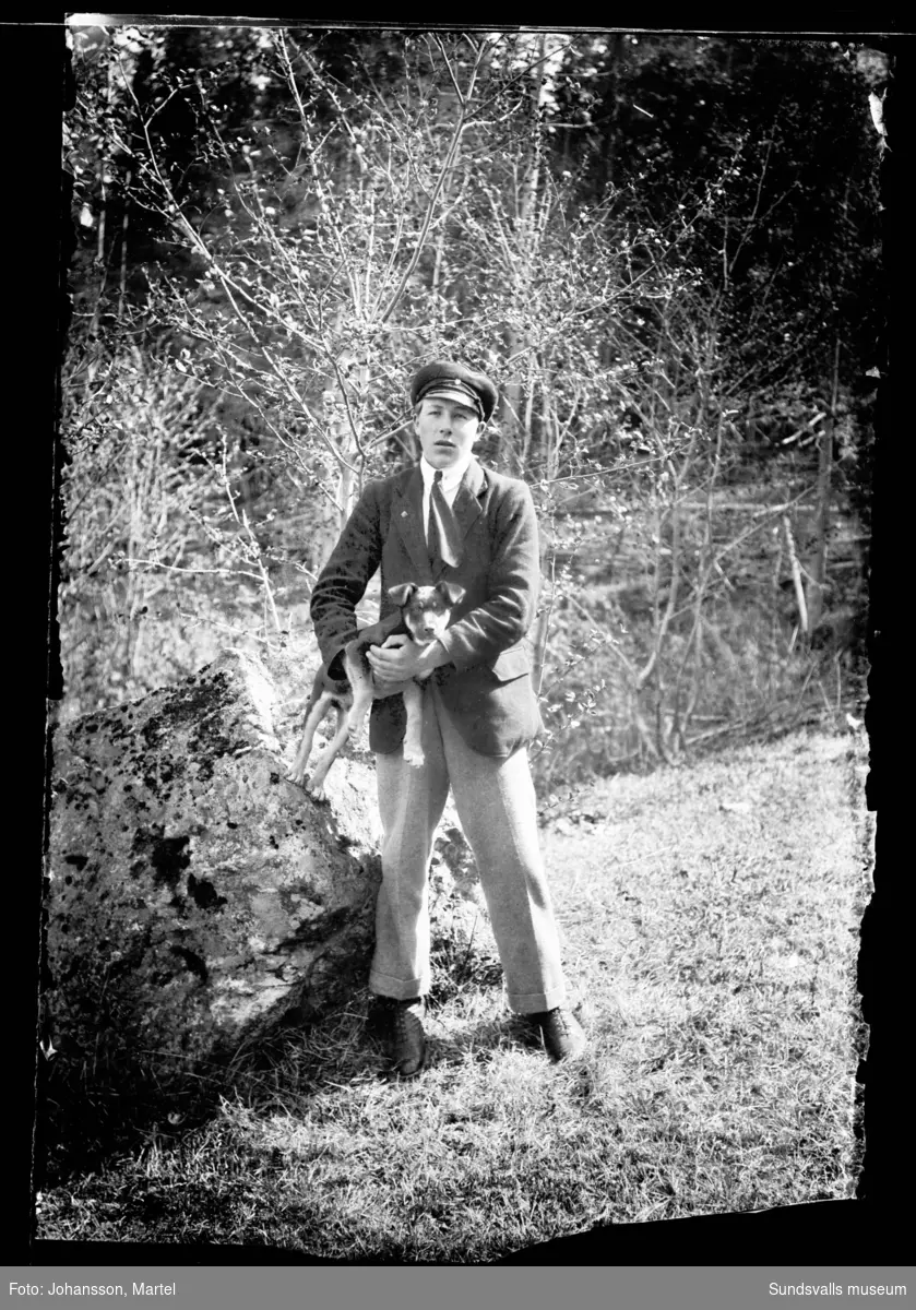 En ung man med skärmmössa och en hundvalp i famnen. Fotograferat i en trädgård.