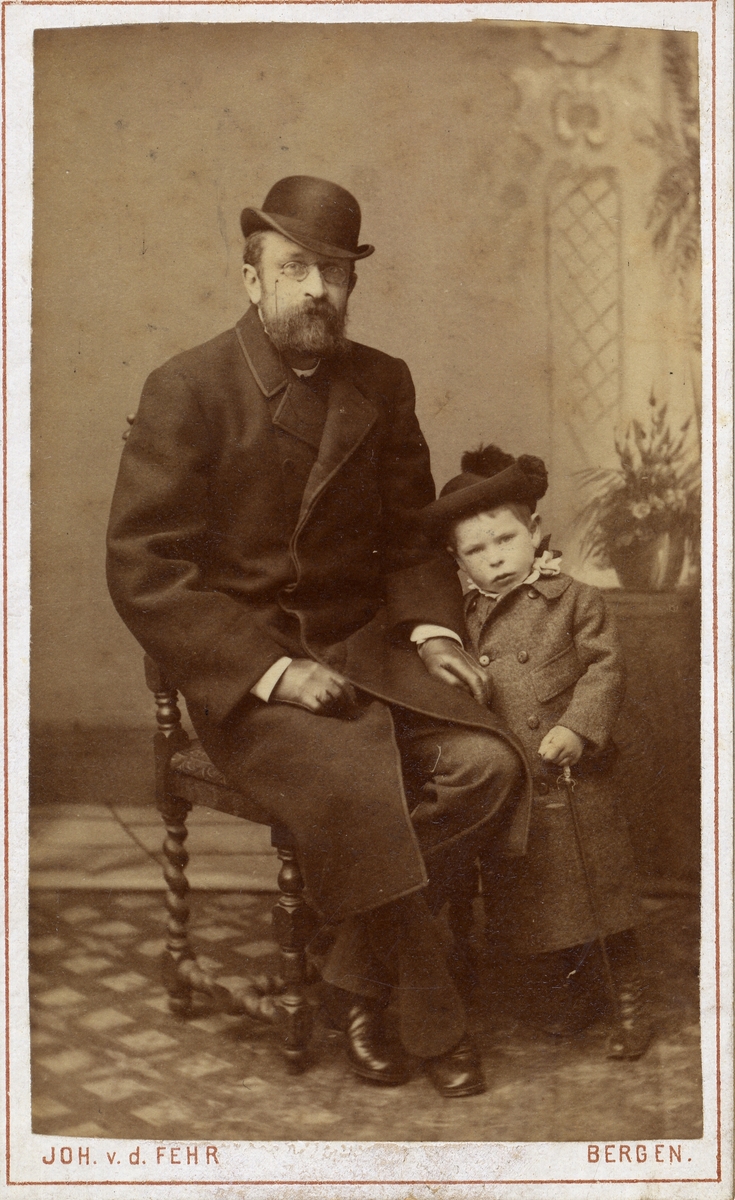 Portrett av sittende mann og liten gutt med hatt og frakk. Far og sønn.