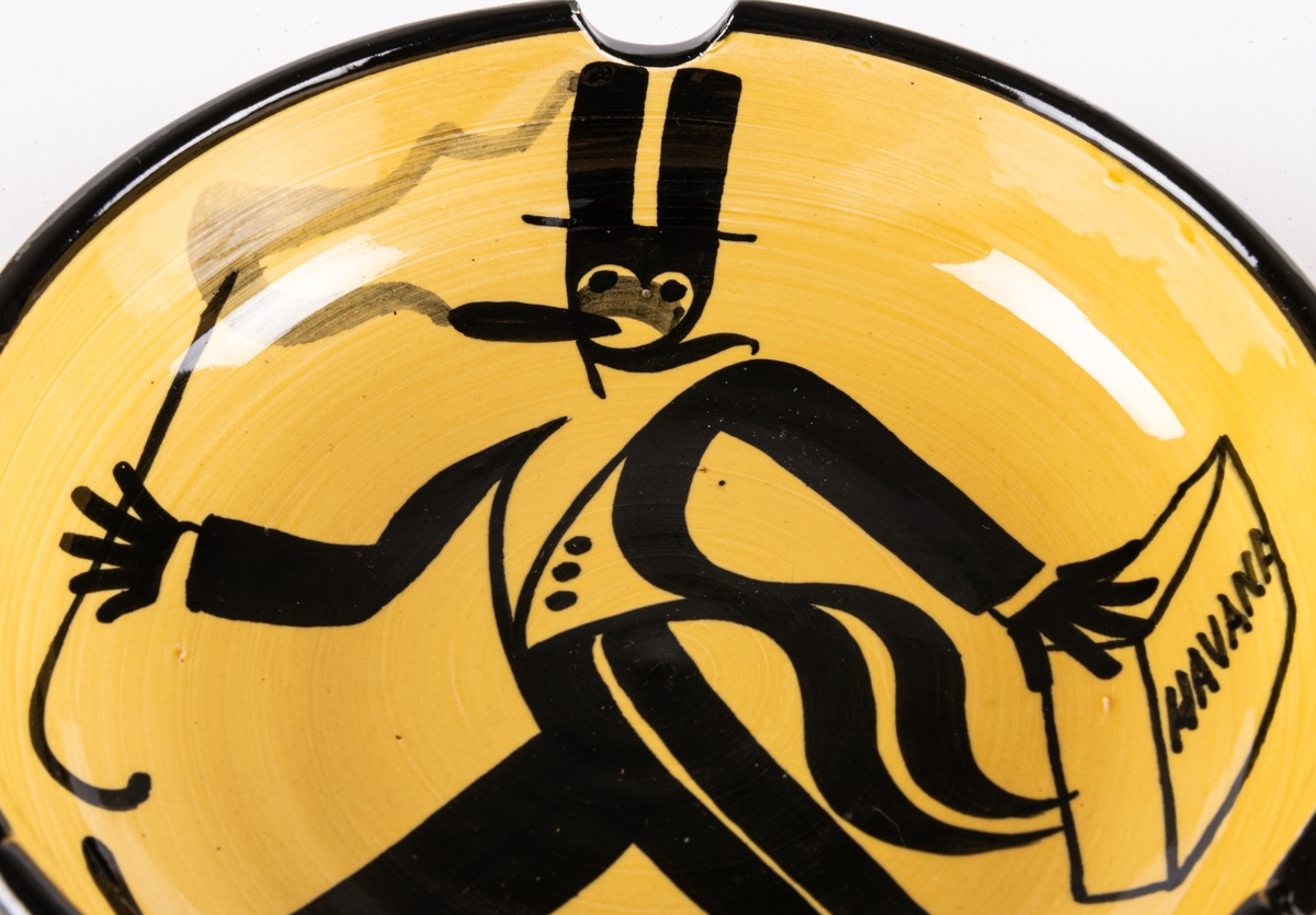 Askfat modell 1081 med cigarrökande herre som motiv och med text "Havanna". Askfatet är tillverkat vid Bo Fajans i Gävle och formgiven av Allan Ebeling. Fatet har gul bottenglasyr med svart handmålad dekor.