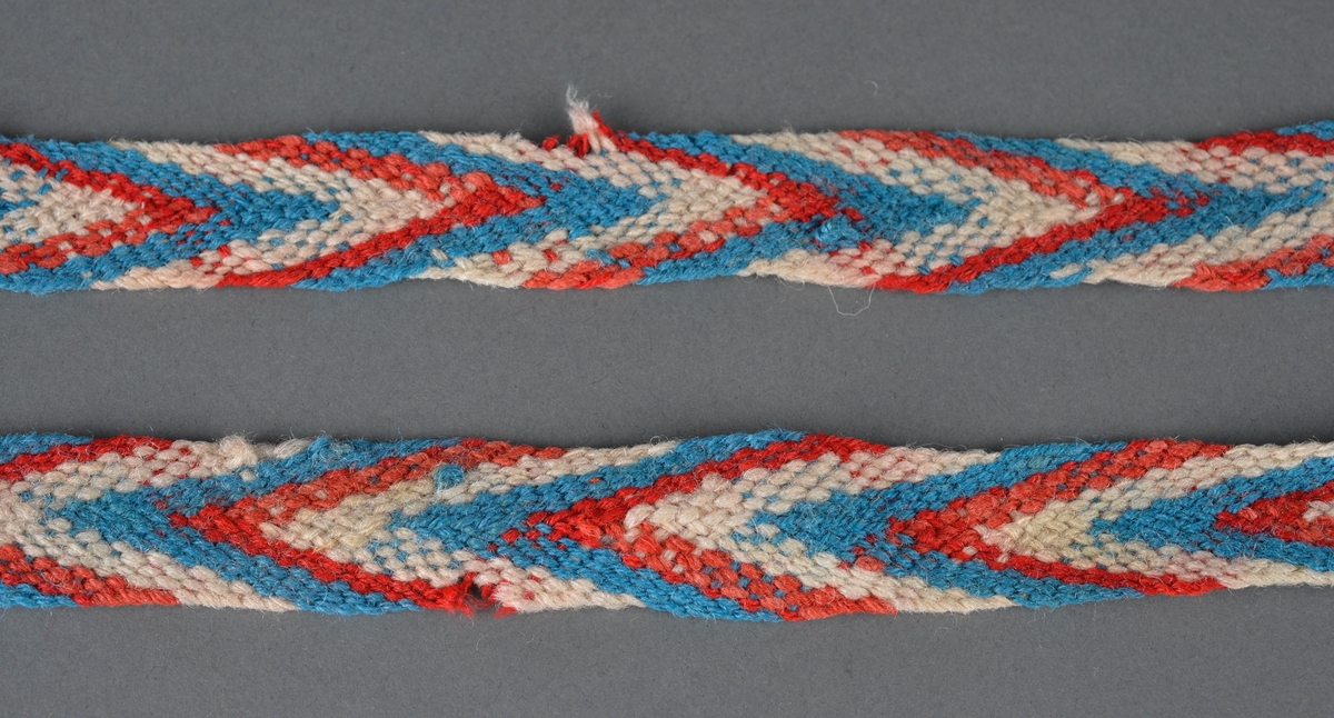 Strømpeband, hoseband fletta i rødt, blått og sauekvitt ullgarn.
Dusk i ene enden. Mangler den andre båndet.
