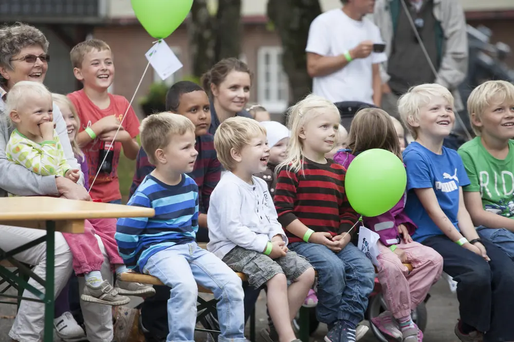 En rekke leende barn sitter på en benk og ser mot noe vi ikke kan se. En har en grønn ballong. En annen sitter på fanget til en bestemor.