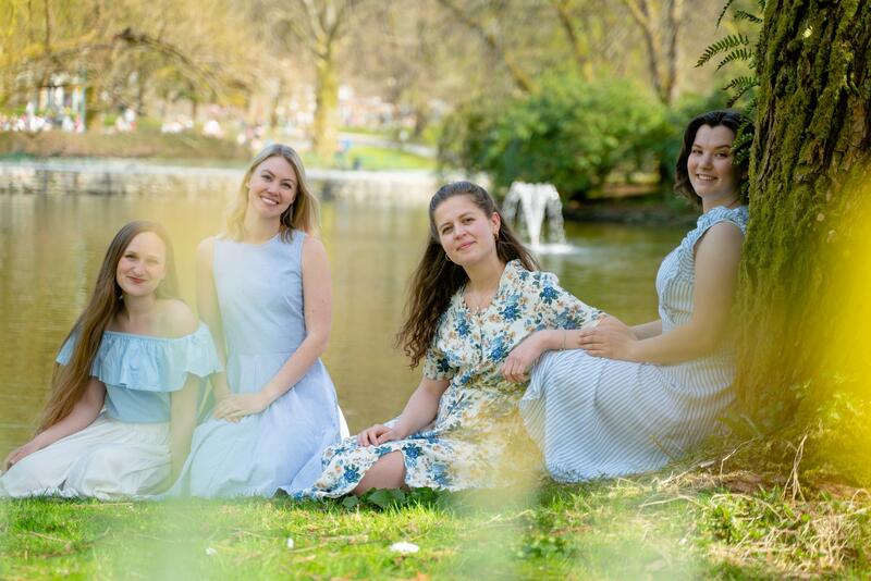 Vokalgruppa Firr er fire unge kvinner som her sitter i hvite, florlette kjoler på grønt gress med et vann i bakgrunnen.