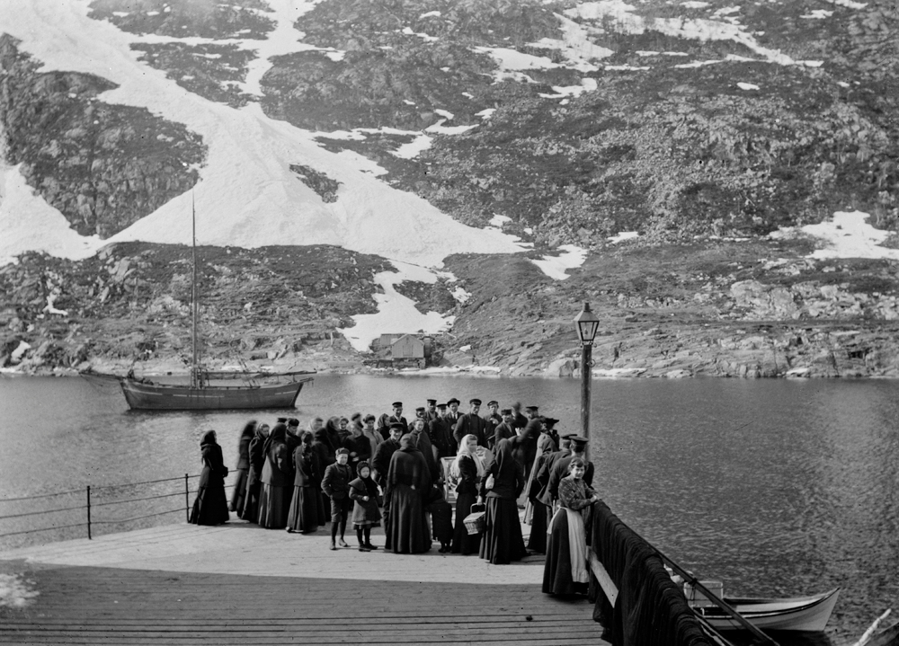 Fra kommunekaiet. Blant folket som venter på fløtmannsbåten er Adolf Schei med slekt og venner. Han reiste til Amerika. Sannsynligvis Edvin Tverå ved stolpen. I Øyfjellet ser vi et mølnhus som tilhørte garver Arntsen.