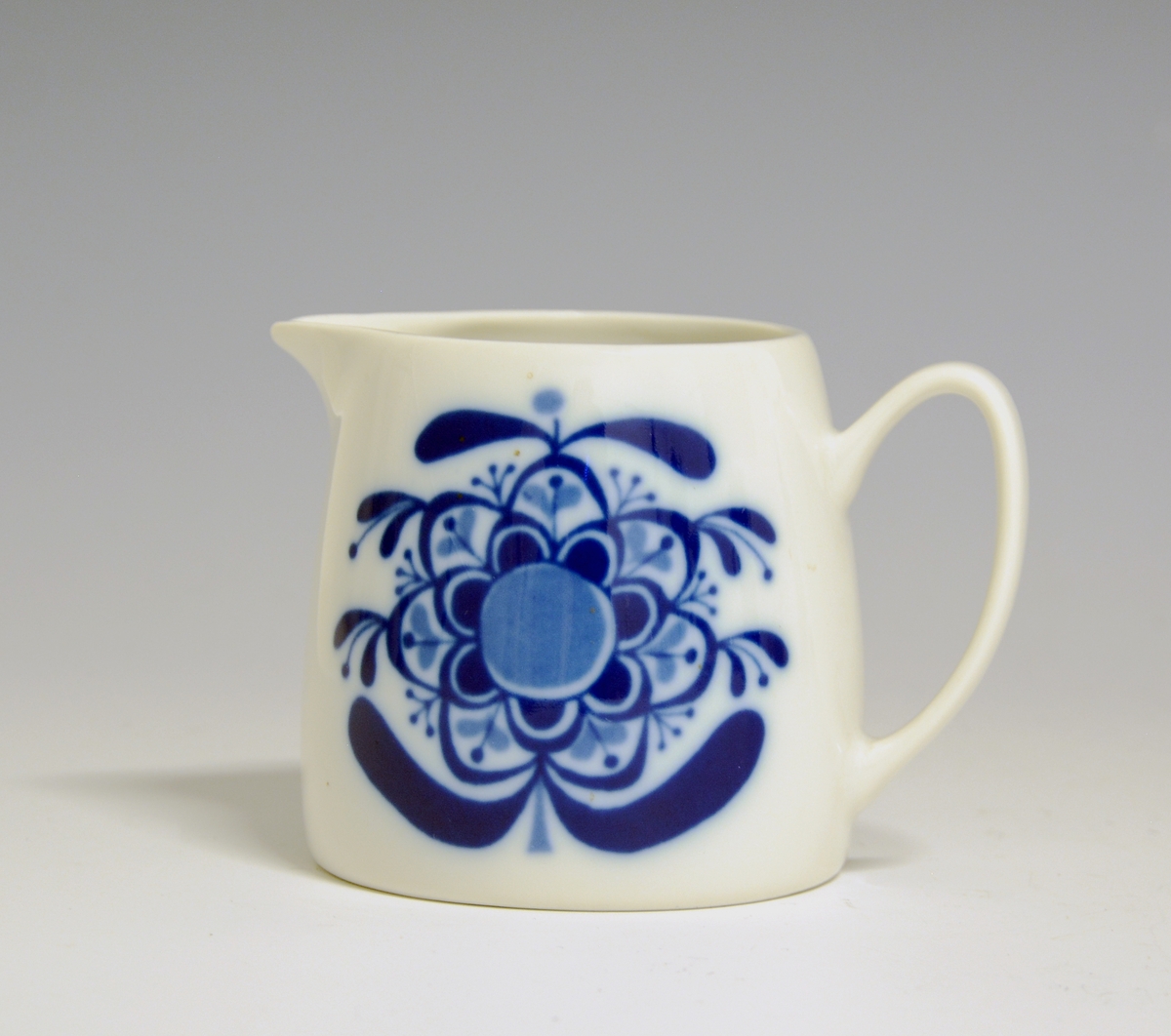 Fløtemugge i brunglasert porselen. Formgitt av Eystein Sandnes. Dekorert med blomsterlignende motiv i blå i-glasurdekor.
Modell: Contrast 2360.
Dekor: Beth Breyen