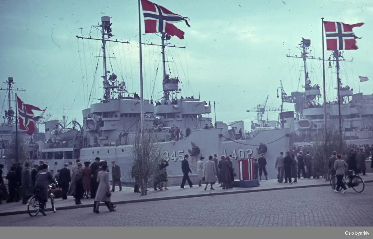 Folk spaserende på Rådhusplassen ved Honnørbrygga under feiringen av frigjøringsdagene 1945, trolig 13.mai. ..Fartøyene på bildet er havgående minesveiperene tilhørende RN 40th Minesweeper Flotilla som ledet styrken inn Oslofjorden. Nærmest HMS Cynthia J345 og HMS Elfreda J402.